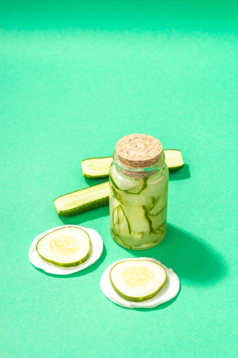 A jar of cucumber kimchi with vibrant red spices, placed on a wooden table surrounded by fresh cucumbers and chili peppers.