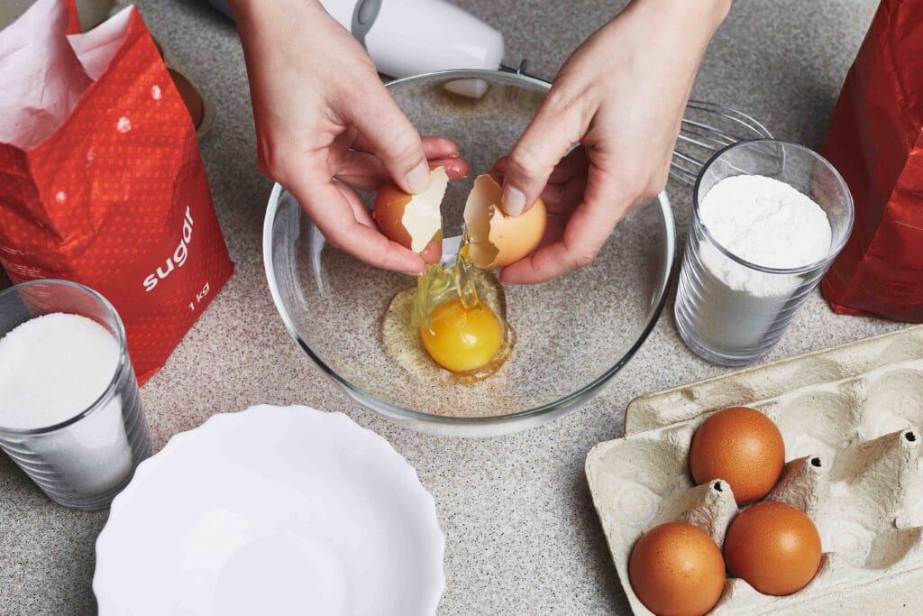 A mixing bowl with batter and a hand holding an egg above it